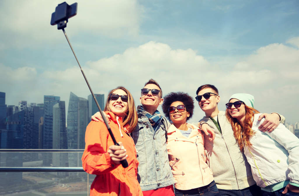 Group of friends taking a selfie using a smartphone on a monopod with a cityscape in the background.